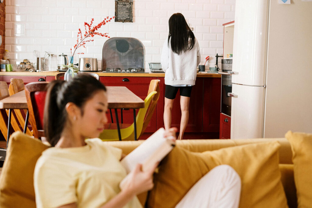 A girl in the foreground reading book on couch while another girl cleans dishes in the background